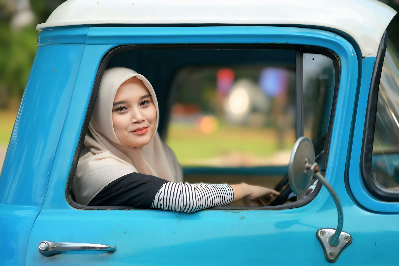 A smiling woman in a hijab sits in a classic blue truck, enjoying a sunny day.