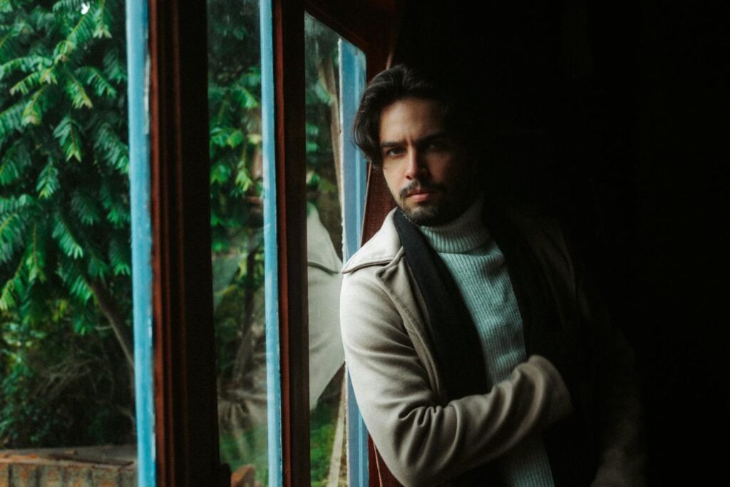 A thoughtful man gazes out a window, wearing a cozy sweater and coat indoors.