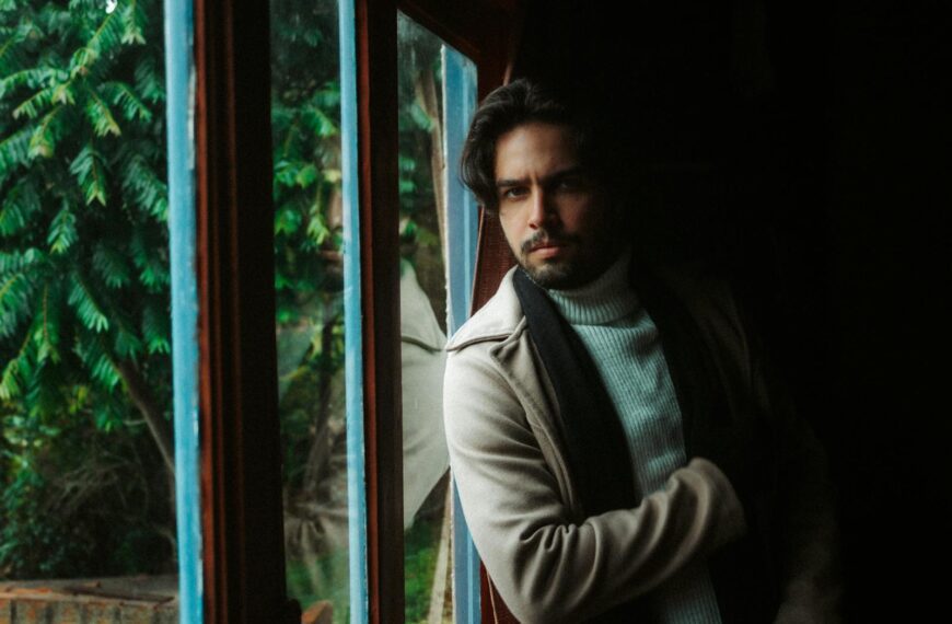 A thoughtful man gazes out a window, wearing a cozy sweater and coat indoors.