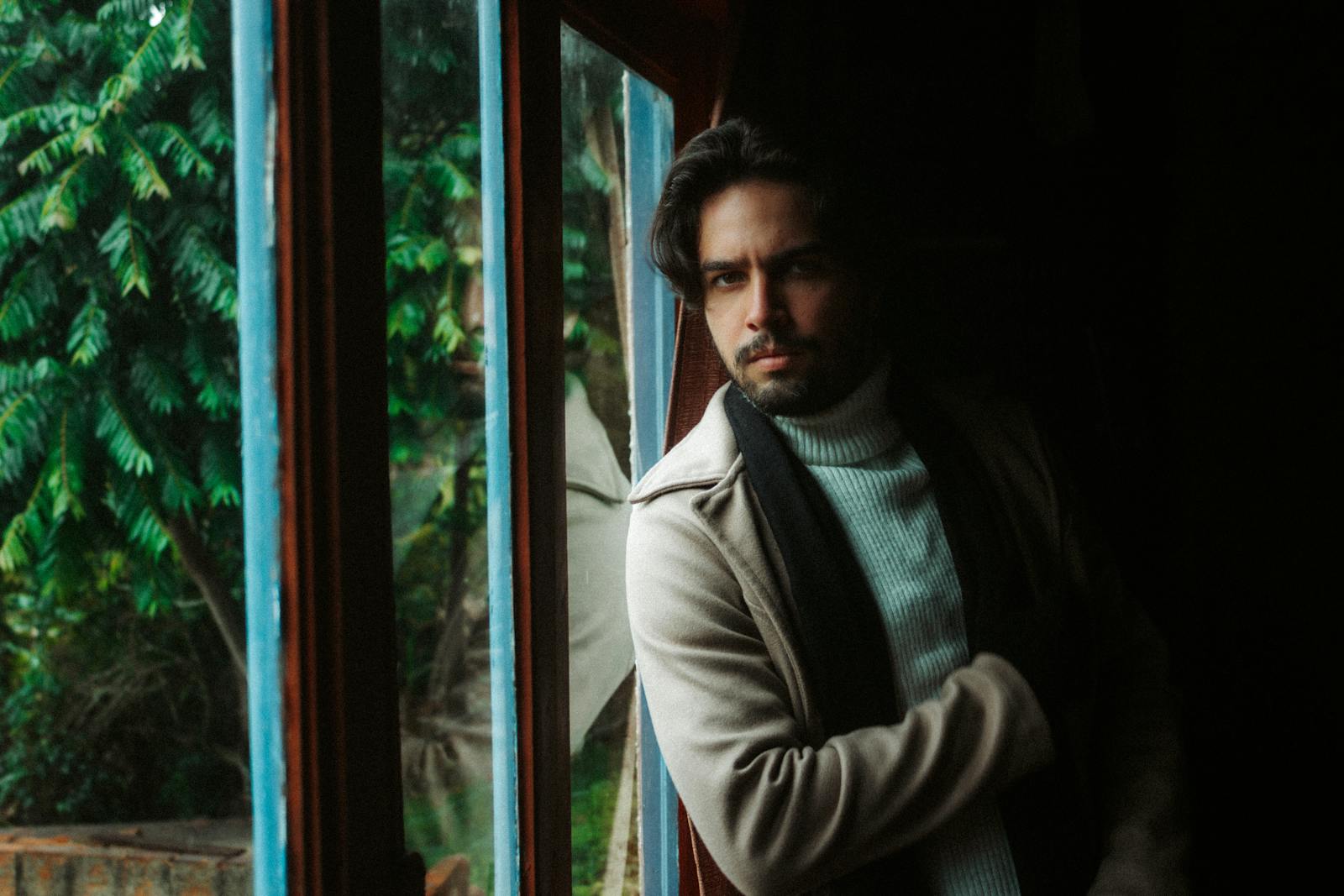 A thoughtful man gazes out a window, wearing a cozy sweater and coat indoors.