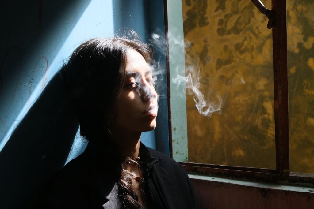 A woman smokes by a window with dramatic lighting casting shadows.