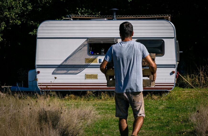 A man carrying a wooden crate approaches a caravan in a sunlit grassy area, evoking a rustic outdoor lifestyle.
