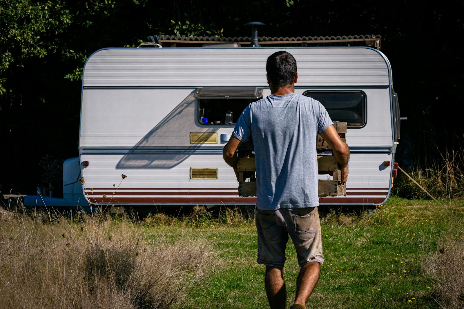 A man carrying a wooden crate approaches a caravan in a sunlit grassy area, evoking a rustic outdoor lifestyle.