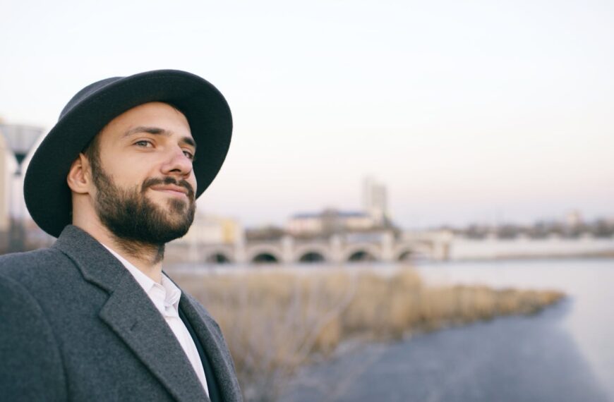 A bearded man in a hat smiling by the river with city skyline in the background.
