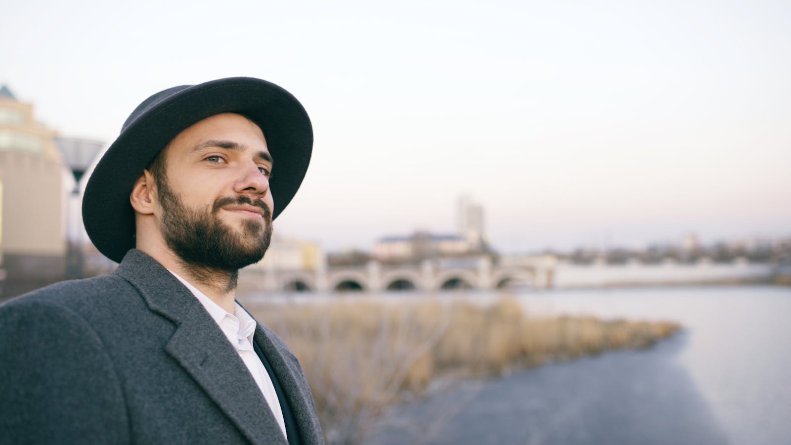 A bearded man in a hat smiling by the river with city skyline in the background.