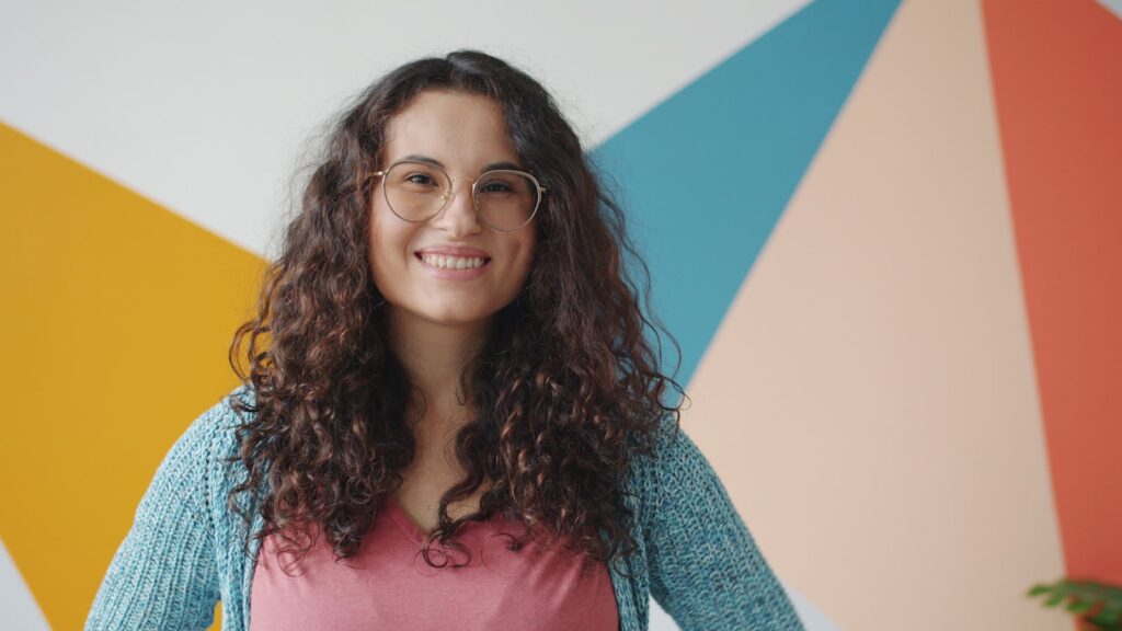 Happy young woman with curly hair and glasses posing indoors. Vibrant colors in the background.