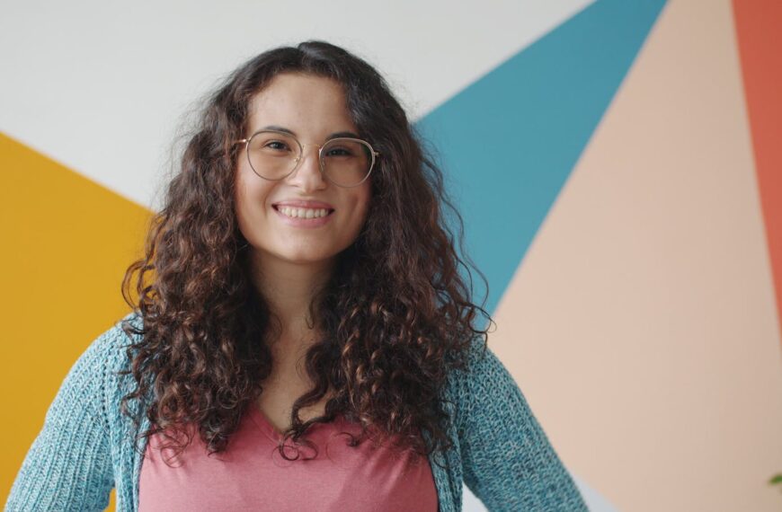 Happy young woman with curly hair and glasses posing indoors. Vibrant colors in the background.