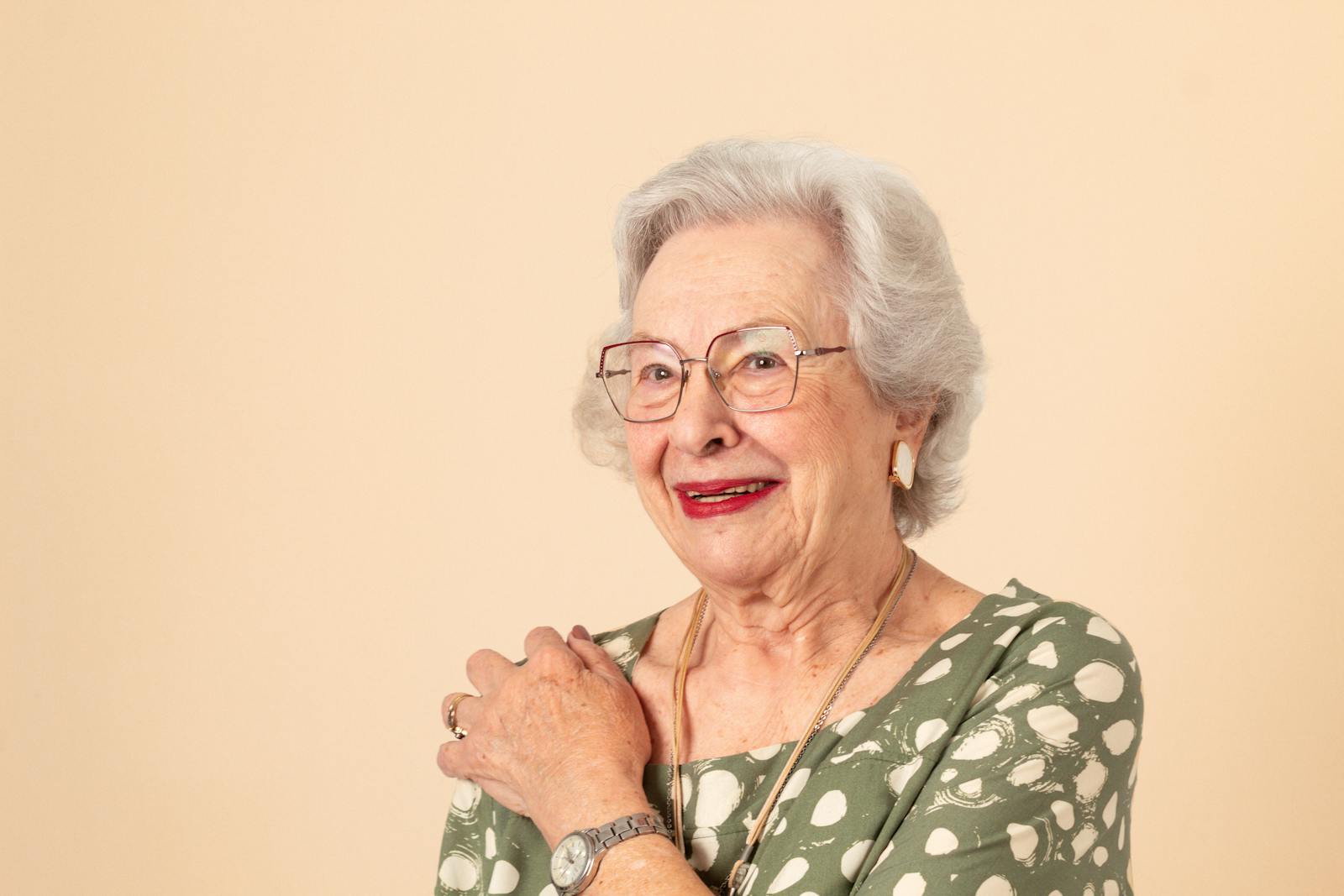 Portrait of a joyful senior woman in a green dress with polka dots, wearing glasses and smiling warmly.