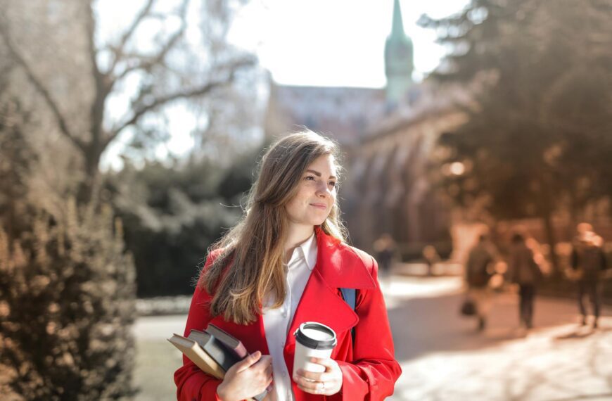 Smiling woman in red coat with coffee and books, enjoying leisure time outdoors on a university campus.