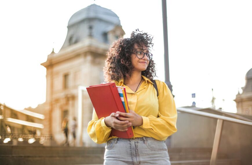 Smiling female student with curly hair and glasses holding books on campus steps.
