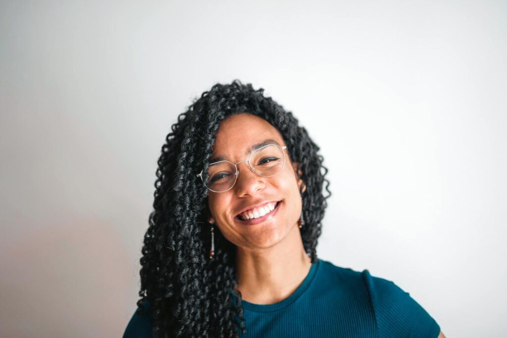 Cheerful young woman with curly hair and glasses smiling in a casual outfit.