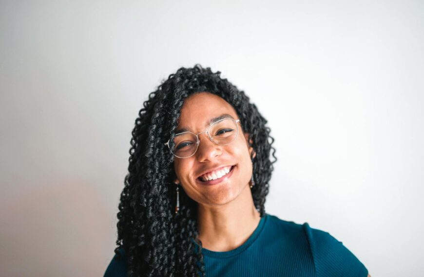Cheerful young woman with curly hair and glasses smiling in a casual outfit.