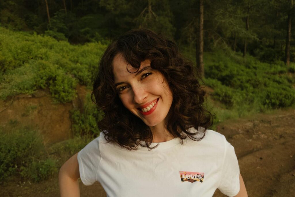 A cheerful woman with curly hair smiling outdoors in a green forest setting.