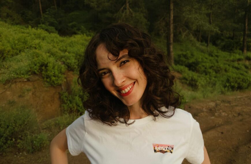 A cheerful woman with curly hair smiling outdoors in a green forest setting.