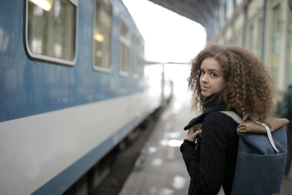 Back view of young student with curly hair in casual warm coat standing on platform with backpack near train and looking at camera over shoulder