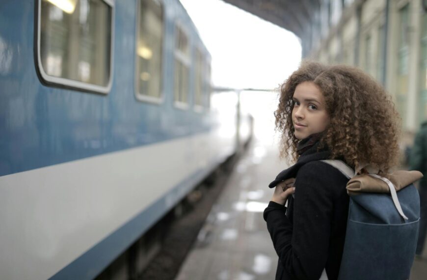 Back view of young student with curly hair in casual warm coat standing on platform with backpack near train and looking at camera over shoulder