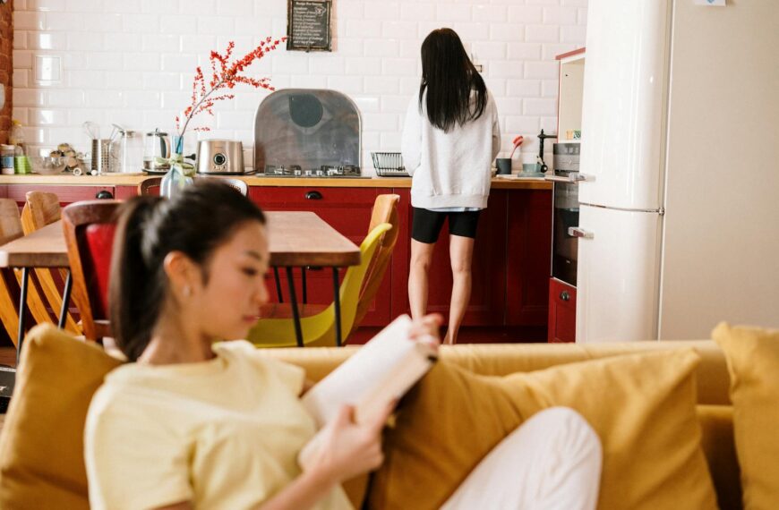 Two women relax at home. One reads on the sofa while the other is at the kitchen counter.