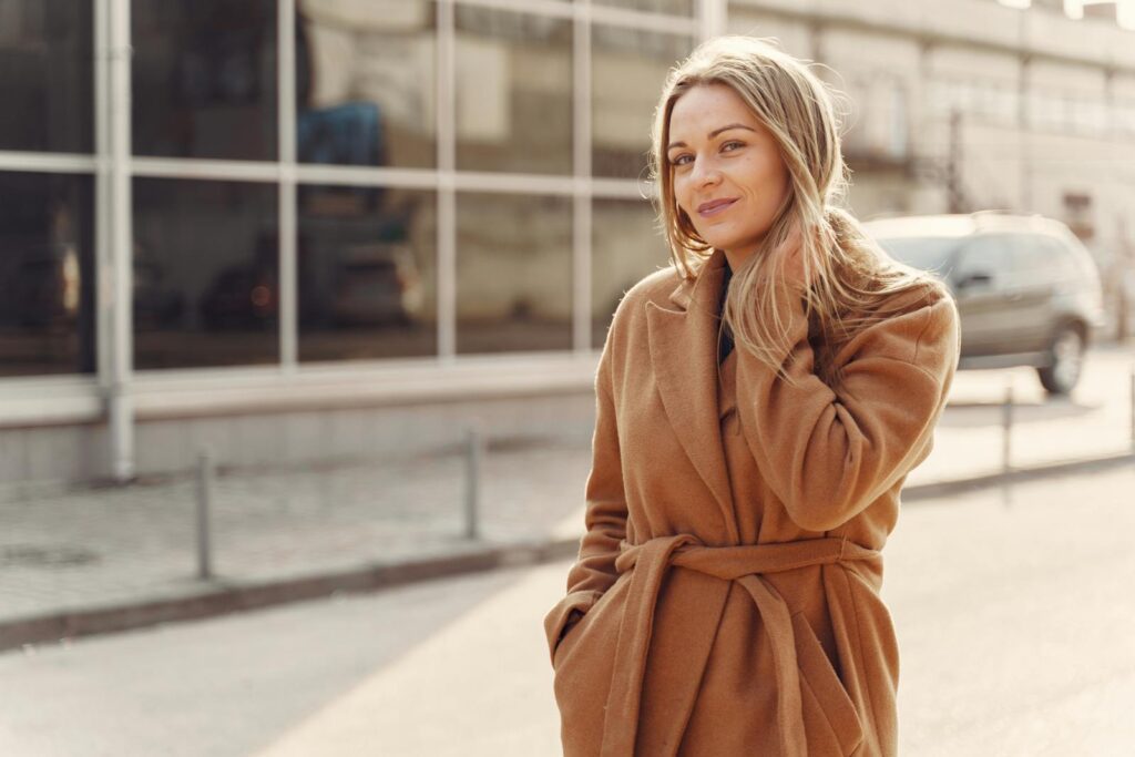 Positive female in stylish warm coat standing on city street against modern glass building and touching hair in back lit