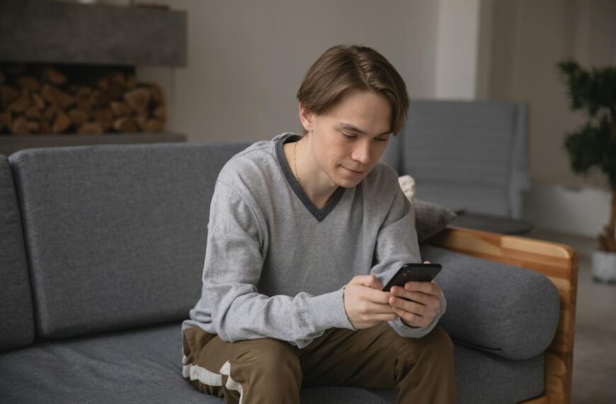 A young man comfortably sitting on a gray couch using his smartphone indoors.