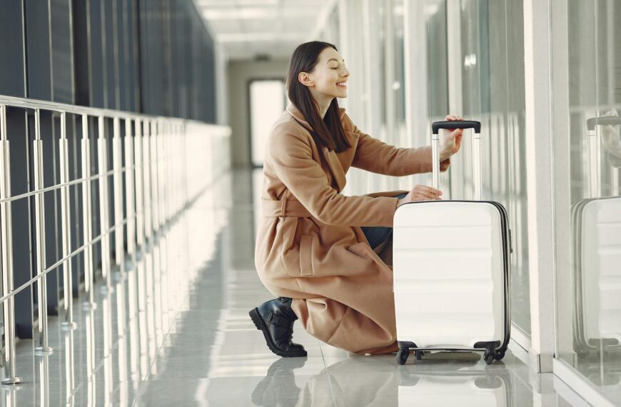 Side view full length of cheerful young female passenger in stylish beige coat with suitcase hunkering down and looking out glass wall in contemporary airport corridor