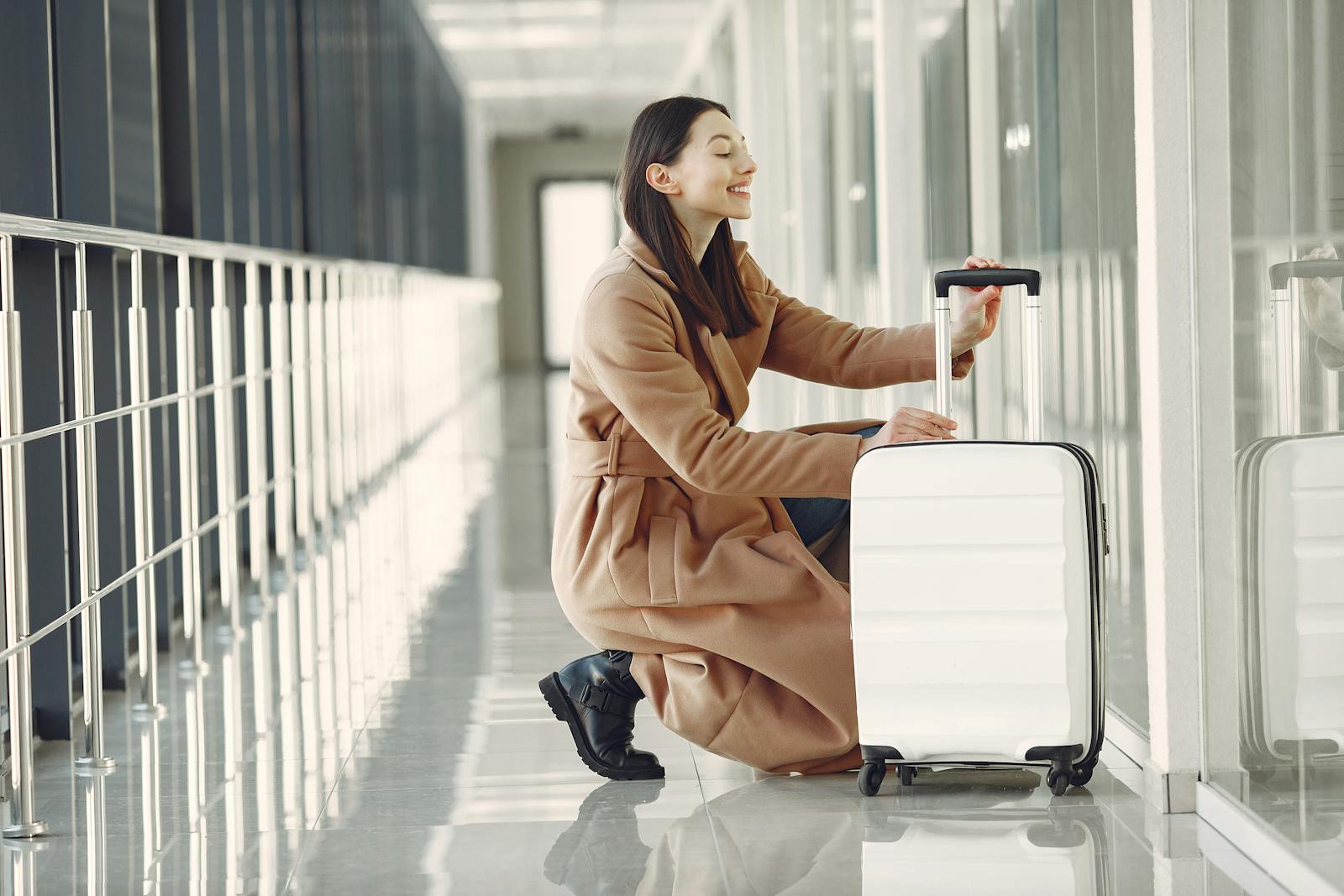 Side view full length of cheerful young female passenger in stylish beige coat with suitcase hunkering down and looking out glass wall in contemporary airport corridor