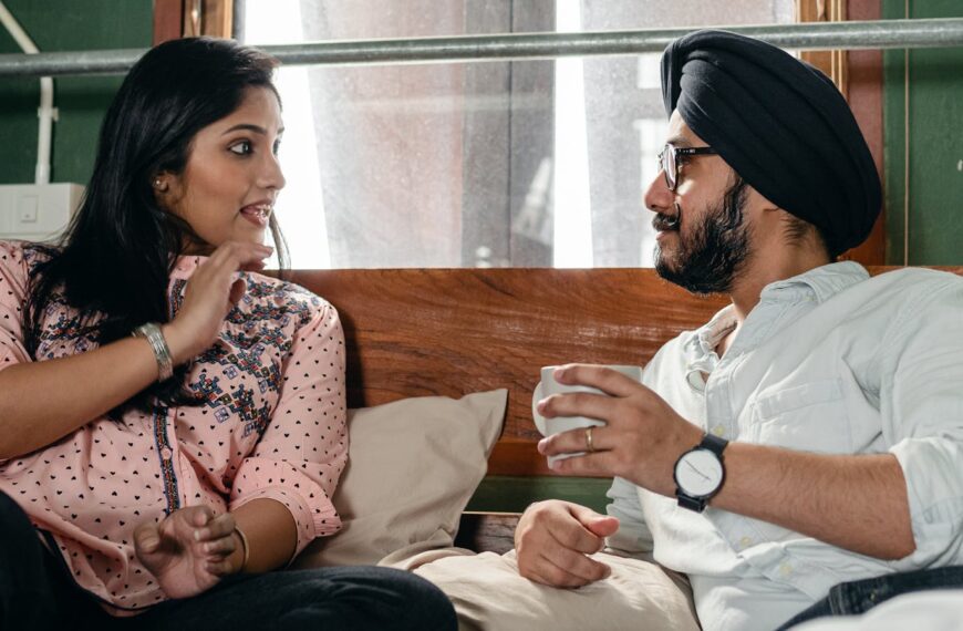 Positive happy Sikh couple wearing casual clothes and turban having lively conversation while resting on comfortable bed with cup of hot drink