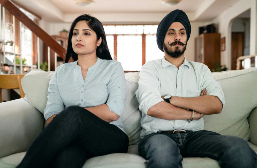 Resentful young Indian couple ignoring each other while sitting on couch together with crossed arms