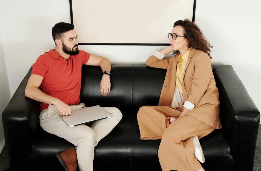 A man and woman sit on a black sofa, having a professional conversation.