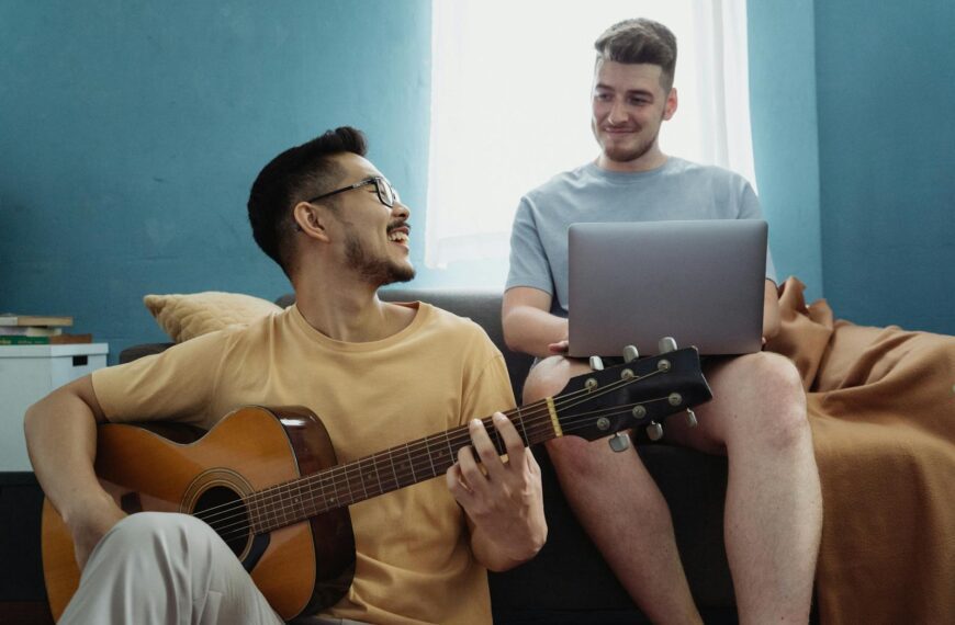 Two friends relax together, one playing guitar and the other using a laptop, creating a joyful atmosphere indoors.
