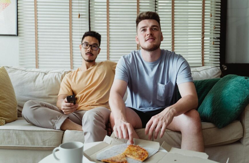 Two friends sitting on a couch, sharing pizza, and watching TV in a comfortable living room.