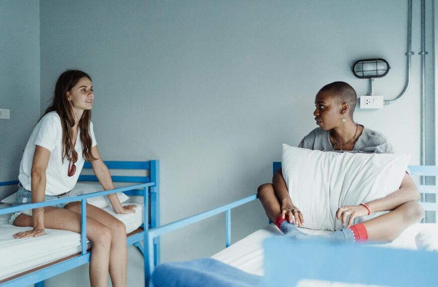 Two women sit and converse casually on bunk beds in a hostel room setting.