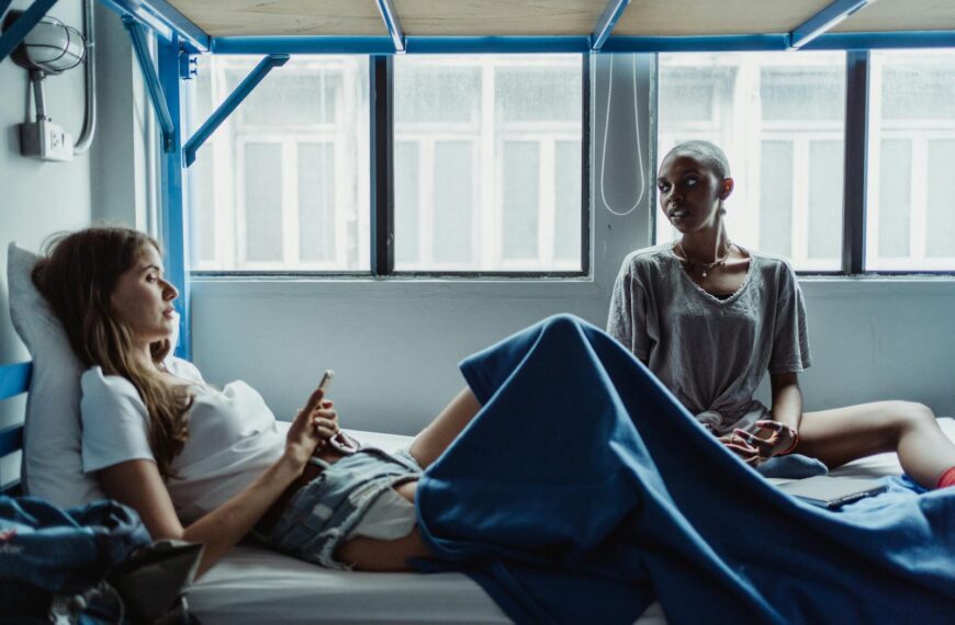 Two women sitting on a bunk bed, talking and relaxing in a hostel room.