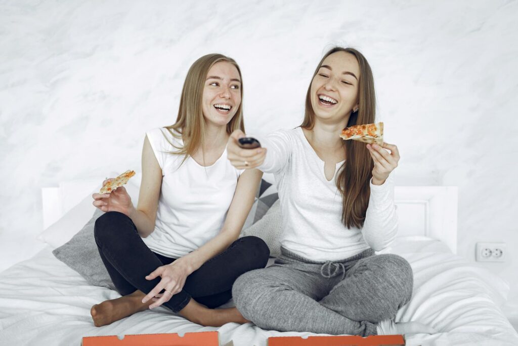 Two young women laughing while eating pizza and watching TV in a cozy indoor setting.