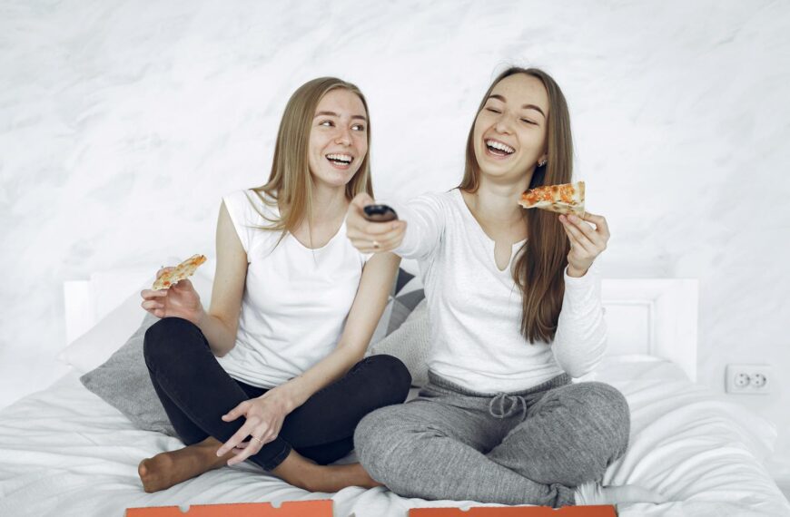 Two young women laughing while eating pizza and watching TV in a cozy indoor setting.