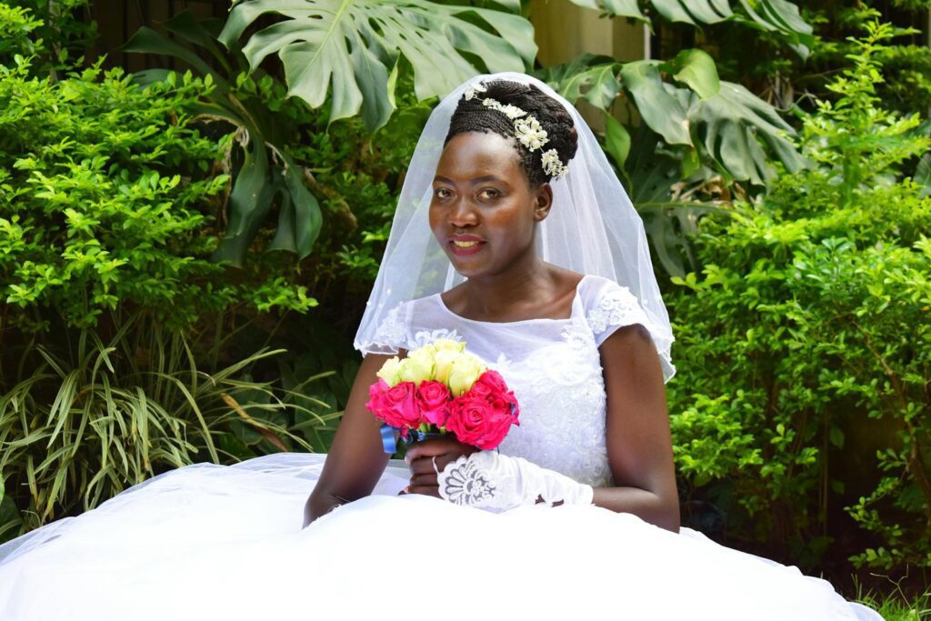 African bride in a white gown and veil holding a vibrant floral bouquet in a lush outdoor setting.