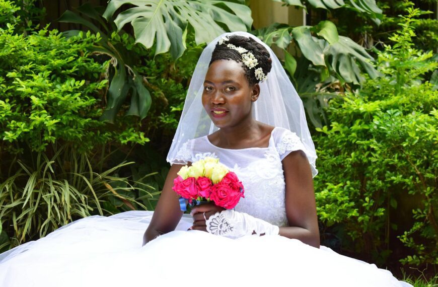 African bride in a white gown and veil holding a vibrant floral bouquet in a lush outdoor setting.