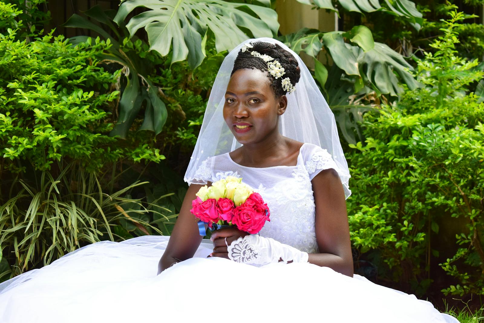 African bride in a white gown and veil holding a vibrant floral bouquet in a lush outdoor setting.
