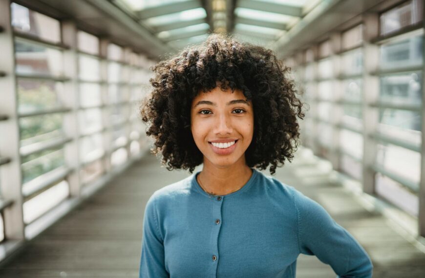 Cheerful African American female with Afro hairstyle smiling and looking at camera while standing on elevated pedestrian crossing with blurred background