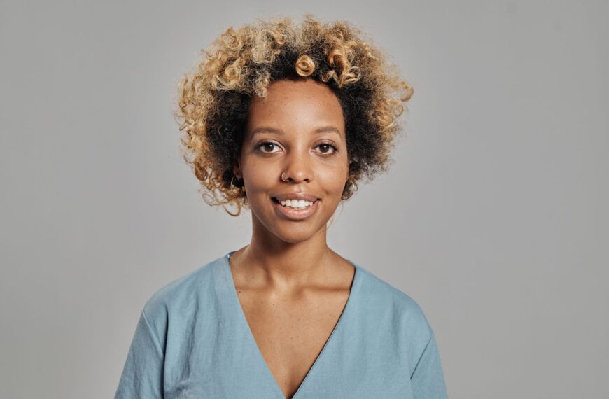 Close-up portrait of a happy woman with curly hair and a nose ring against a gray background.