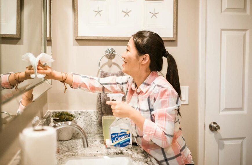 An adult woman cleaning a bathroom mirror using a spray bottle and cloth.