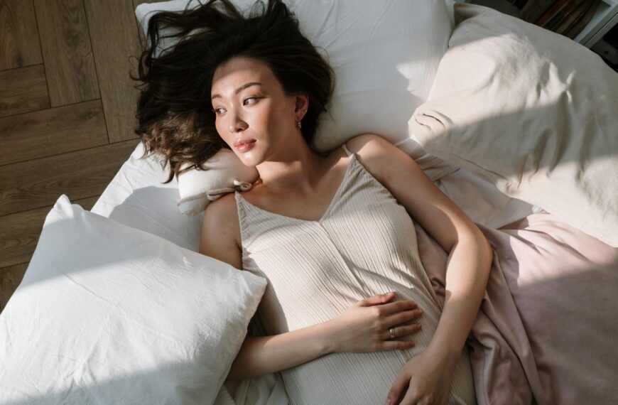 Asian woman in sleepwear lying on bed with hand on belly, enjoying morning sunlight.