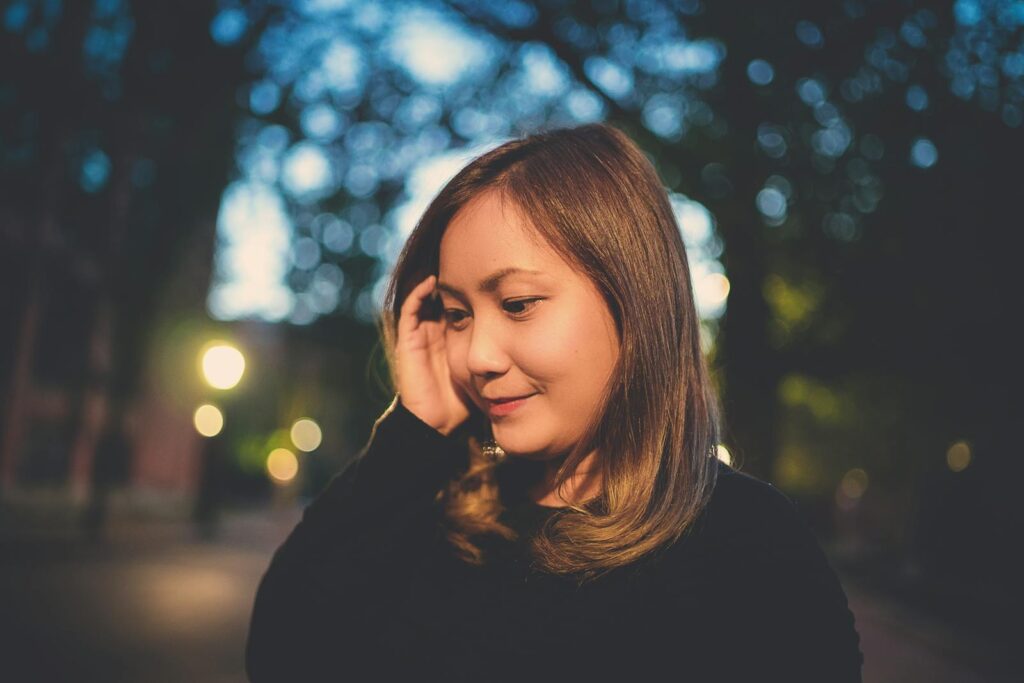 Calm Asian woman in casual clothes touching hair and thoughtfully looking away on street in evening time