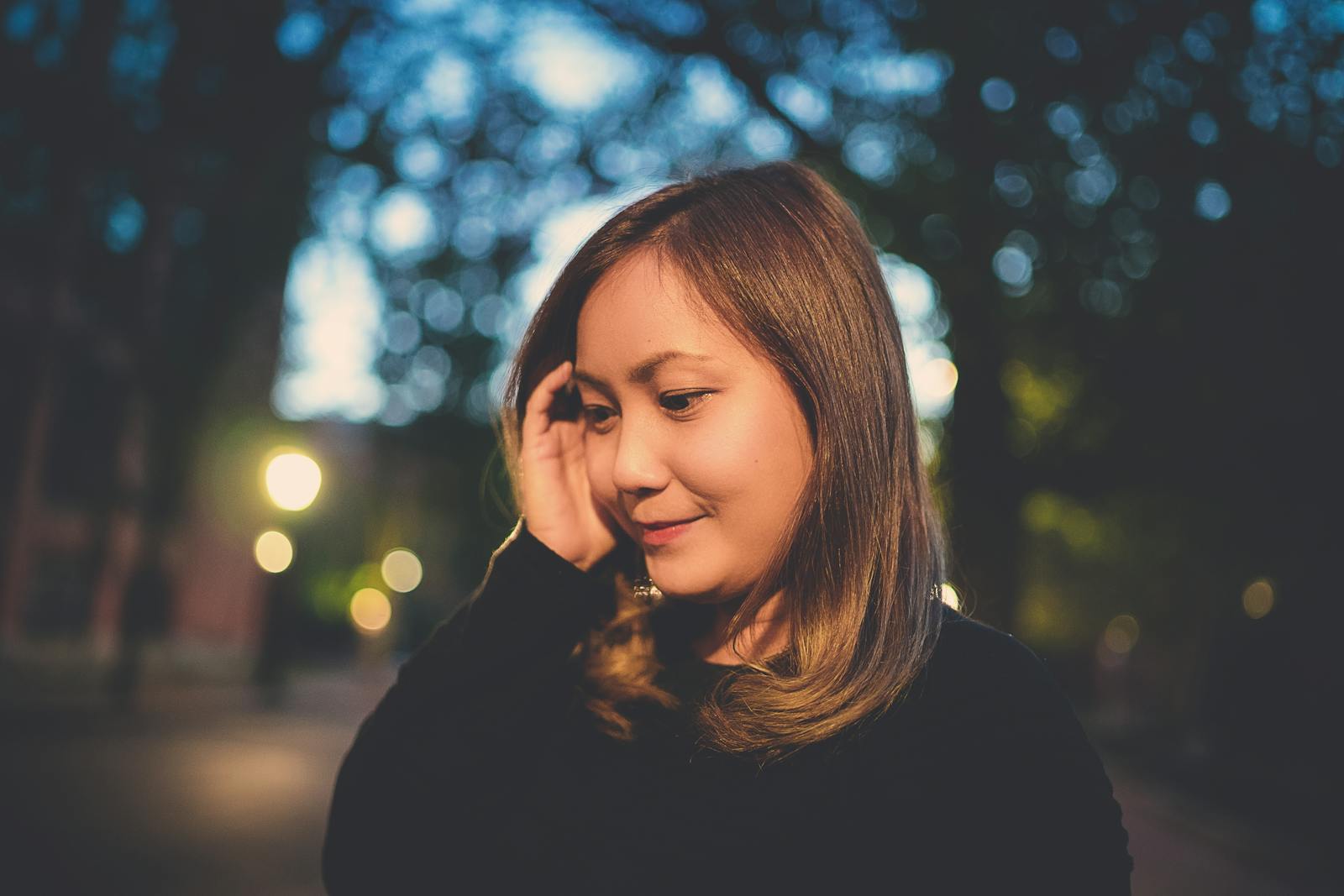 Calm Asian woman in casual clothes touching hair and thoughtfully looking away on street in evening time