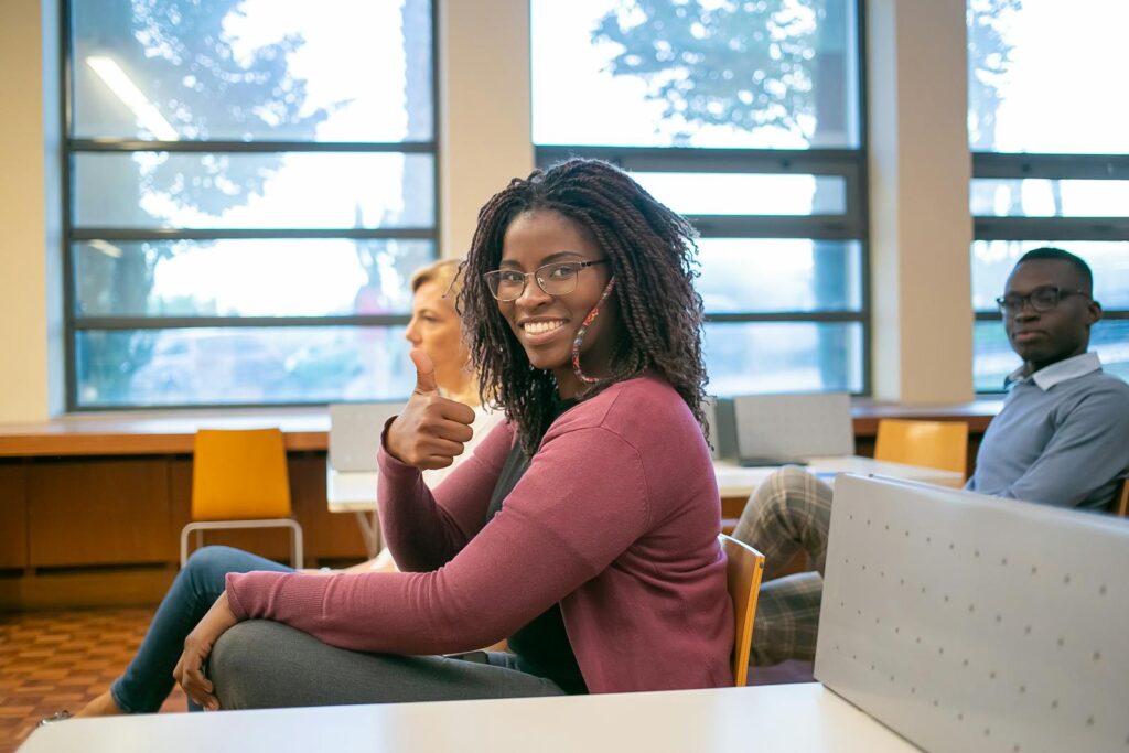 Side view of positive African American female student demonstrating like sign while sitting at lecture with classmate and teacher
