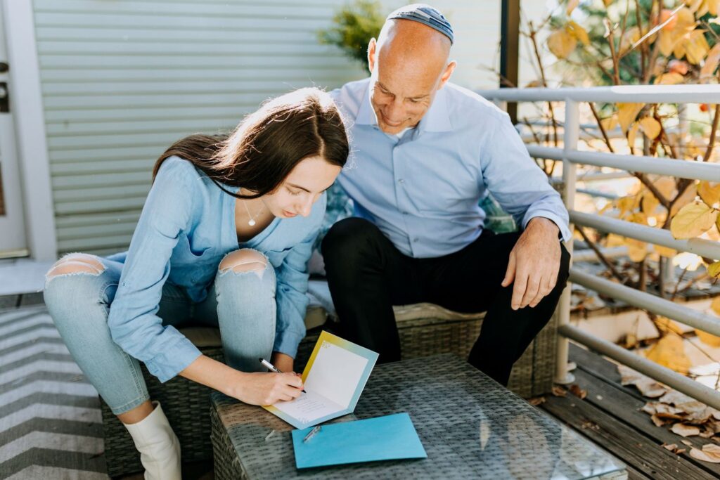 A father and daughter are sitting outdoors, writing Hanukkah cards together, enjoying a peaceful moment.