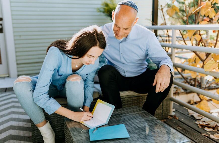 A father and daughter are sitting outdoors, writing Hanukkah cards together, enjoying a peaceful moment.