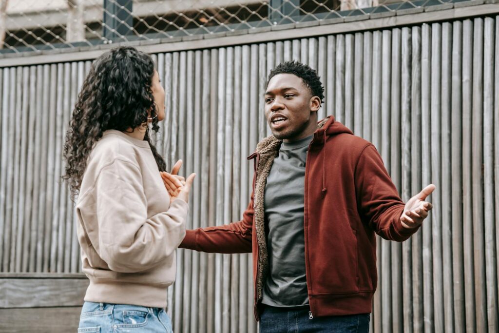 Angry African American boyfriend and ethnic girlfriend looking at each other while having emotional dispute on street near wooden fence