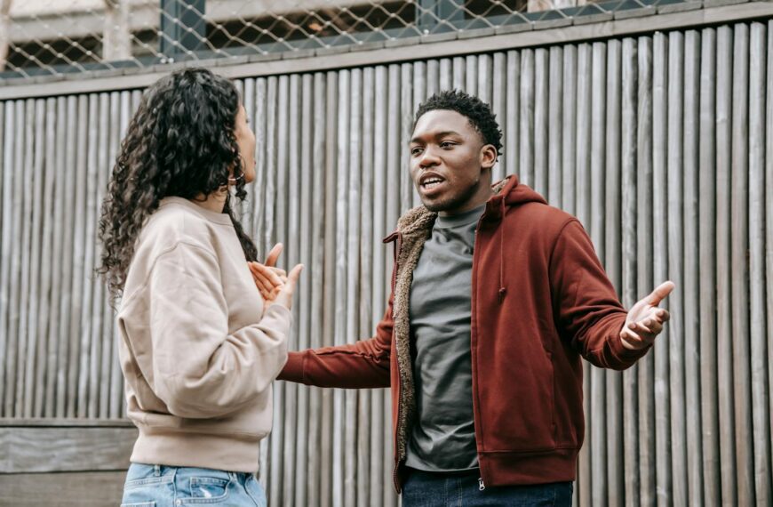 Angry African American boyfriend and ethnic girlfriend looking at each other while having emotional dispute on street near wooden fence