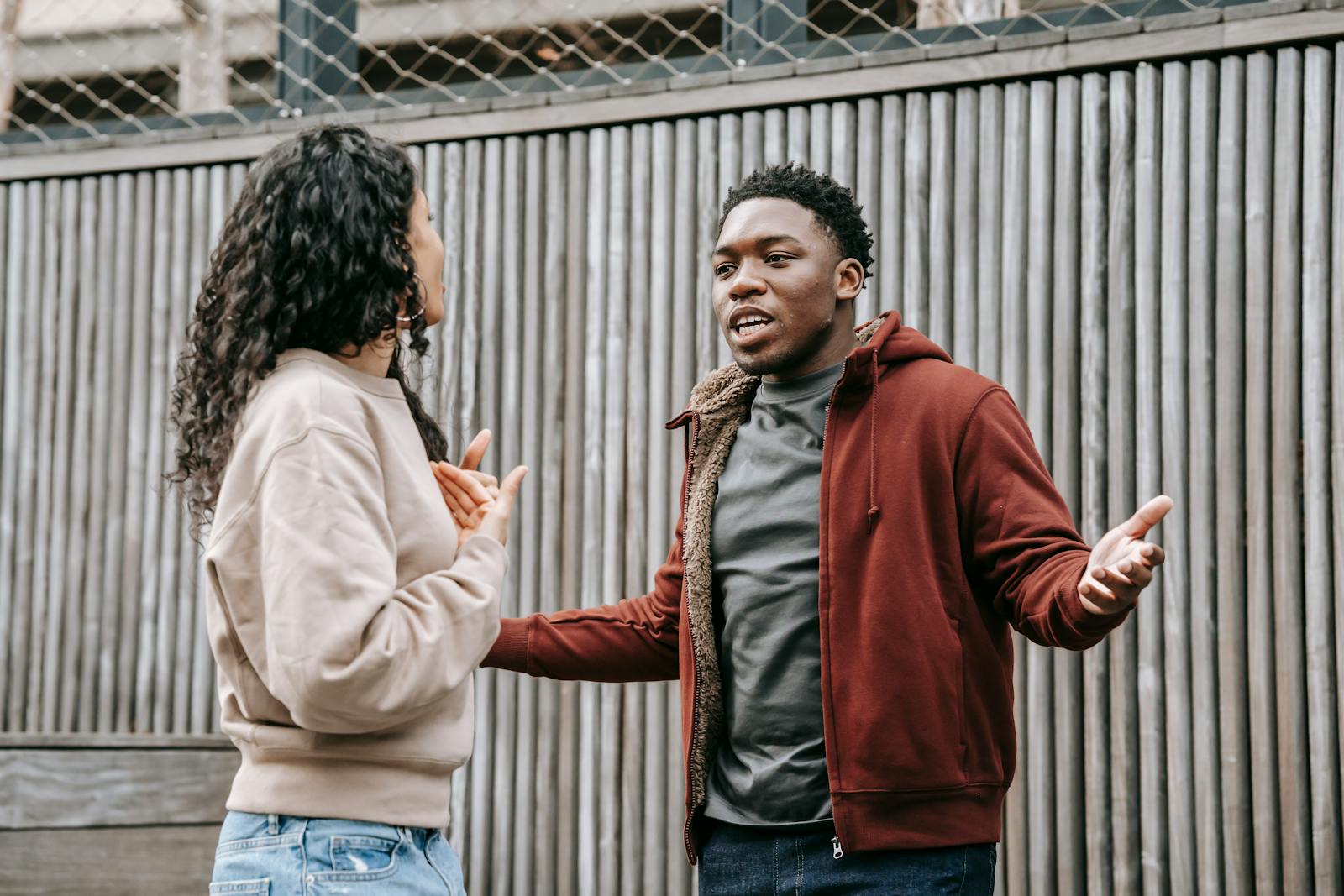 Angry African American boyfriend and ethnic girlfriend looking at each other while having emotional dispute on street near wooden fence