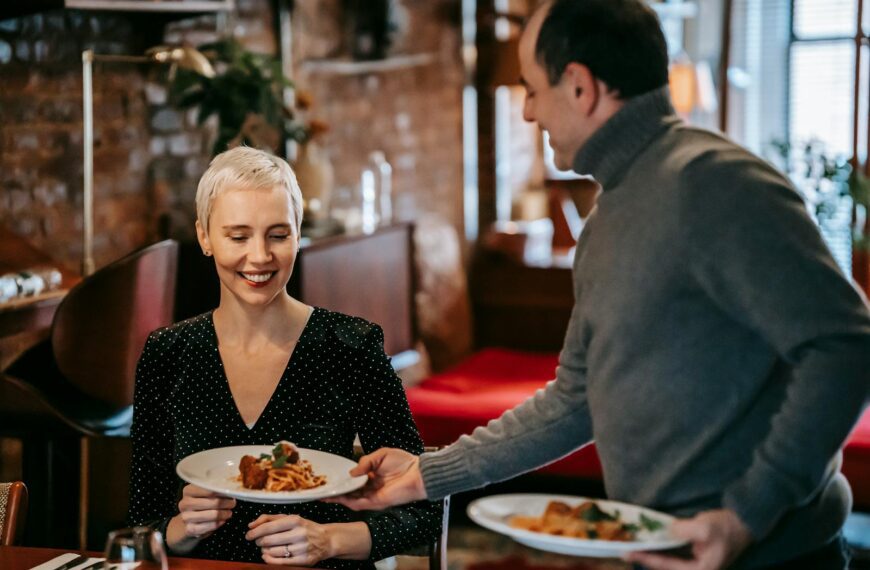 Positive adult couple having dinner in bright room near table while man serving plates with tasty pasta with meatballs near chairs in daylight